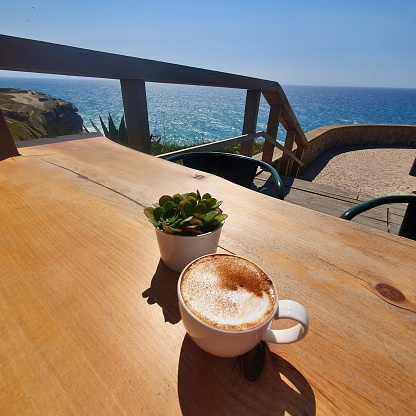 Cup of cappuccino and pot of succulent on the wooden table with two metal chairs in the cafeteria on the ocean or sea coastline with water of Atlantic in the background, rocks of cliff