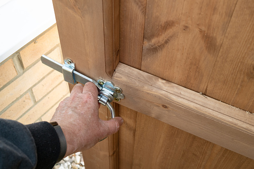 Shallow focus of a homeowner opening a metal latch of a new installed wooden garden gate. The gate leads to an enclosed garden.