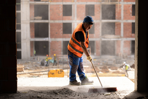 Unrecognizable mid-adult construction worker, in orange colored reflective vest, using broom, to clean the dust on the construction site