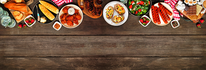 Summer BBQ food top border over a dark wood banner background. Assorted grilled meats, potatoes, vegetable dishes and smores platter. Overhead view.