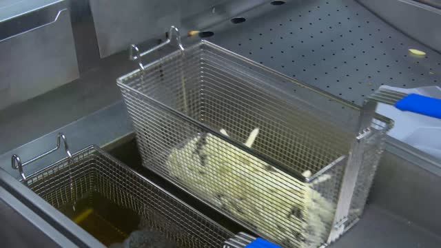 Frying fresh or frozen french fries in a professional looking frying basket submerging by bubbling, boiling hot frying oil. Static camera.