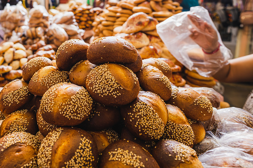 This horizontal photograph offers a close-up view of a selection of artisanal breads displayed at a market in Oaxaca City. Stacks of golden loaves dusted with sesame seeds fill the foreground, their appetizing texture wonderfully captured. In the blurred background, the hand of a Latina woman is discernible as she attends to the bread stand, adding a human touch to the image.