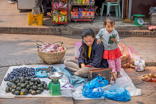 Morning market, Luang Prabang, Laos - March 17th 2023: Female greengrocer with her daughter in her market stall on the ground at the famous morning food market in Luang Prabang which used to be the capital on Laos