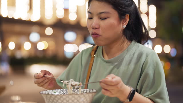 Front view of Asian black long hair woman eating pho noodel in a bowl or paper dinner box night lifestyle near street downtown in Athens Greece with blur bokeh street light background