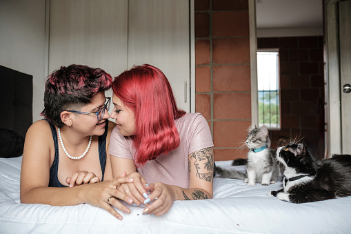 a couple of lesbian women lying in bed, looking at each other with love and tenderness while their cats keep them company. Connection between them is evident in their affectionate glances and gestures. Cats, also part of your family, add extra dose of calm and companionship to this intimate moment.