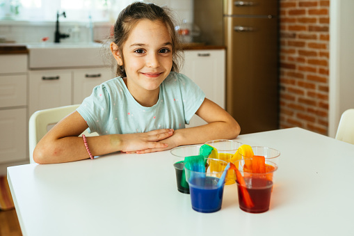 Little girl sitting at the table and watching rainbow food color experiment with water and paper at home