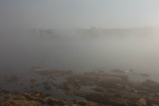 The landscape, the ocean shore in the fog, the fog rises above the stones and closes the view of the city. Atmospheric mystical mood.
