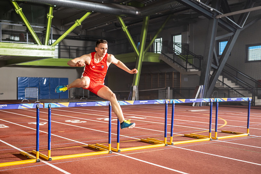 Male caucasian athlete running fast and jumping through hurdle while training at stadium track front side view wide shot