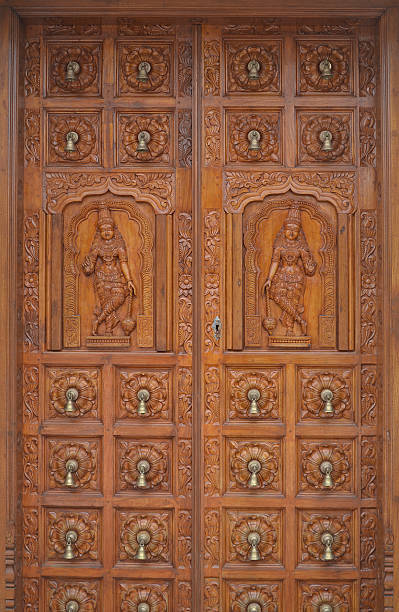 Traditional hindu wood carvings on large double timber door. The doors are studded with small brass bells arranged in a grid pattern. Symmetrical female figures on each leaf of the doors on the middle.