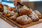 Cropped image of an Asian woman using tongs to pick up an almond croissant from a bakery store
