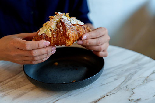 An Asian woman sitting at a table, enjoying her breakfast almond croissant during the morning