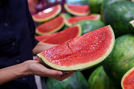 Young Asian woman is grocery shopping for fresh, organic fruits in the supermarket. She is specifically choosing a red and juicy watermelon from the produce aisle. This is part of her routine grocery shopping, as she maintains a healthy eating lifestyle.