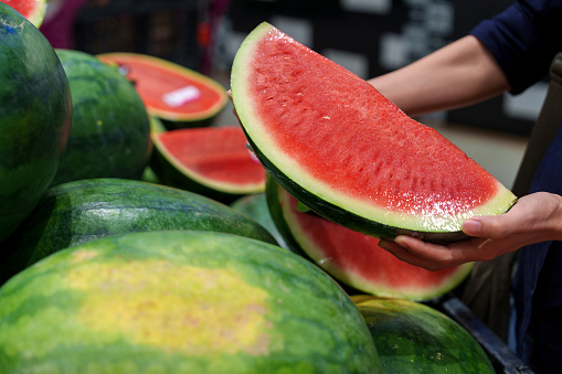 Young Asian woman is grocery shopping for fresh, organic fruits in the supermarket. She is specifically choosing a red and juicy watermelon from the produce aisle. This is part of her routine grocery shopping, as she maintains a healthy eating lifestyle.