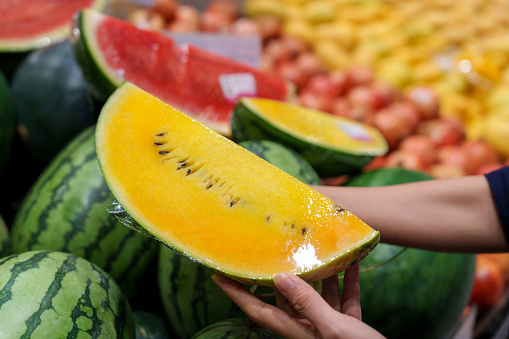 Asian woman is grocery shopping for fresh, organic fruits in the supermarket. She is specifically choosing a yellow and juicy watermelon from the produce aisle. This is part of her routine grocery shopping, as she maintains a healthy eating lifestyle.