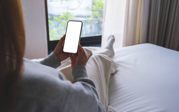 istock Mockup image of woman holding mobile phone with blank desktop white screen while sitting on a bed 1493896426