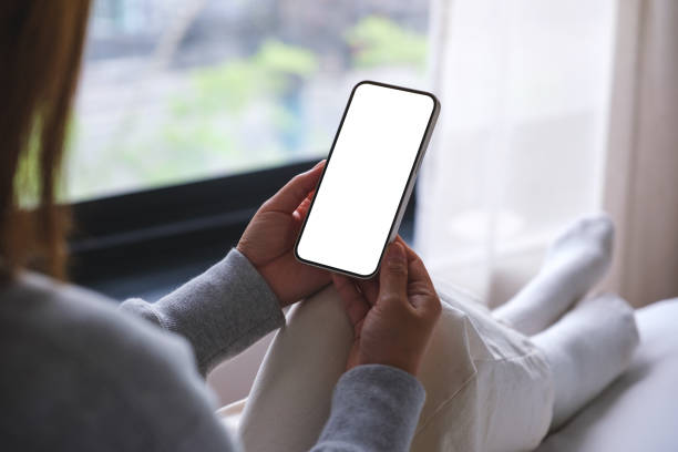 istock Mockup image of woman holding mobile phone with blank desktop white screen while sitting on a bed 1493896428