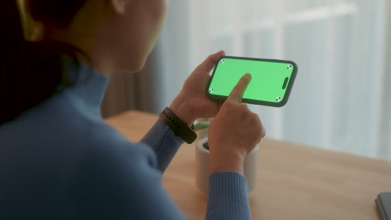 Woman holding smart phone horizontal orientation and tapping with green screen at home office.