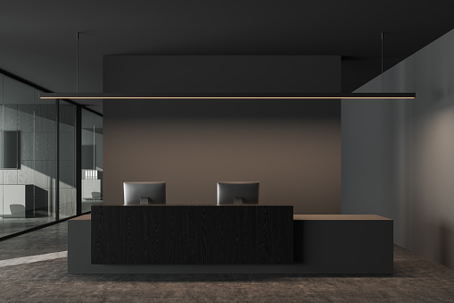 Dark office room interior with reception desk and pc computers, business hallway in company and grey concrete floor. Meeting and coworking space behind glass doors. 3D rendering