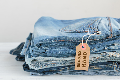 Stack of blue jeans and tag with inscription second hand on white background. Second hand clothing shop. Circular fashion, eco friendly sustainable shopping, thrift stores concept.