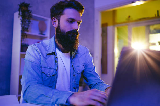 istock Portrait of a skilled young bearded junior front-end developer sitting at a purple and blue-lighted home office and working on software. 1493945094