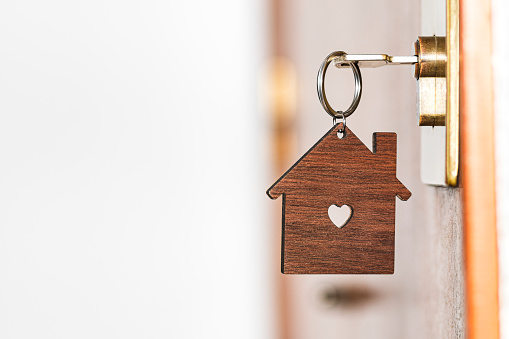 Wooden key chain in the shape of a house with a heart-shaped hole in the center hanging from a key inserted in the lock of a front door of a house.