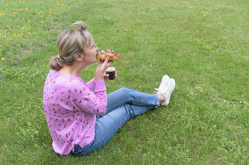 A young woman with a croissant and coffee eats sitting on the grass in the park. Place for text