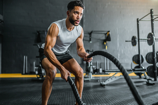 Fit young man focused on training with battle ropes in gym, looking into distance