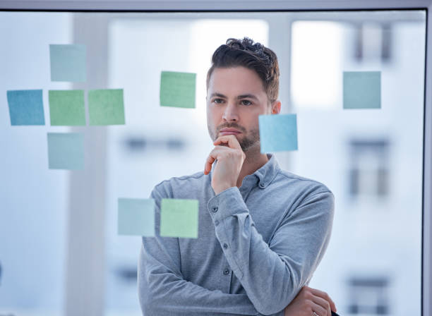 istock Creative man, thinking and brainstorming in schedule planning glass board for tasks or reminder at office. Male wondering in thought for project plan, idea or sticky note strategy for company startup 1493963707