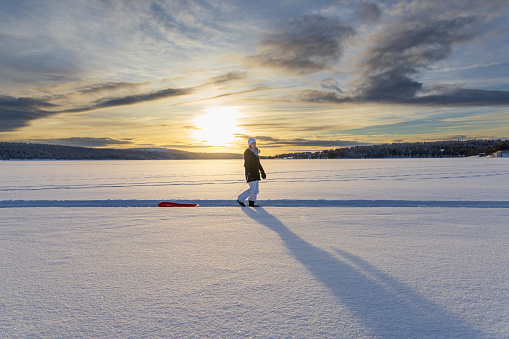 Girl walking over a frozen snow covered lake with a sled during sunset in Lapland, Finland