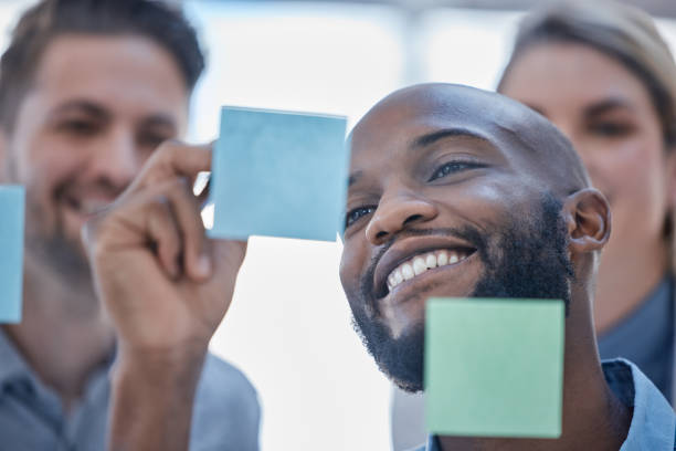 istock Black man, writing and smile for schedule planning on glass board for team brainstorming or tasks at office. Happy African male smiling in project plan, ideas or sticky note for teamwork strategy 1493963734
