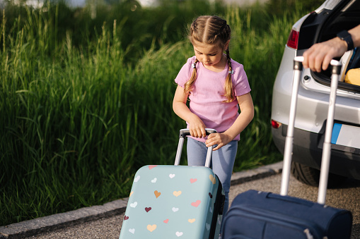 A little girl carries her suitcase to the car. Dad helps his daughter put the luggage in the trunk. High quality photo