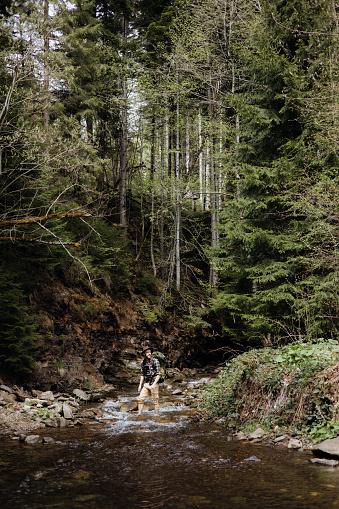 A dynamic action shot capturing the man with a backpack carefully navigating across a mountain stream during hiking. The composition emphasizes his balance and agility as he crosses the flowing water, showcasing the challenges and excitement of outdoor adventure.