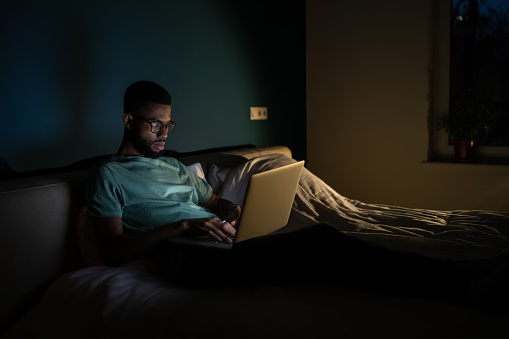 Focused African American man wearing glasses looking at laptop screen working on project until night, lying in bed with laptop. Black guy freelance IT developer coding at night to meet deadline