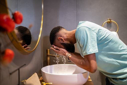Skin care routine for men. Side view of African American millennial guy cleaning skin in morning, standing above bathroom sink and washing face with water, trying to wake up