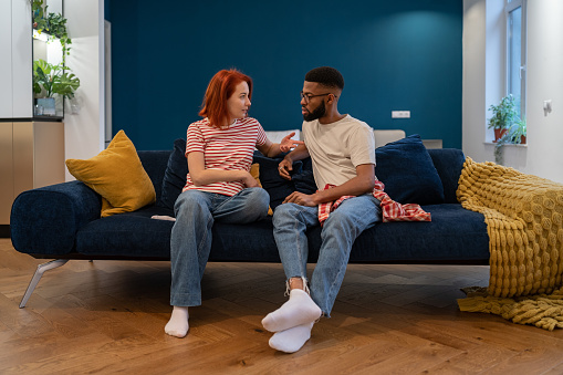 Emotional multi ethnic annoyed stressed couple sitting on couch arguing at home. Man and woman talking face to face trying come to agreement. Misunderstanding, relationship crisis, problems concept.