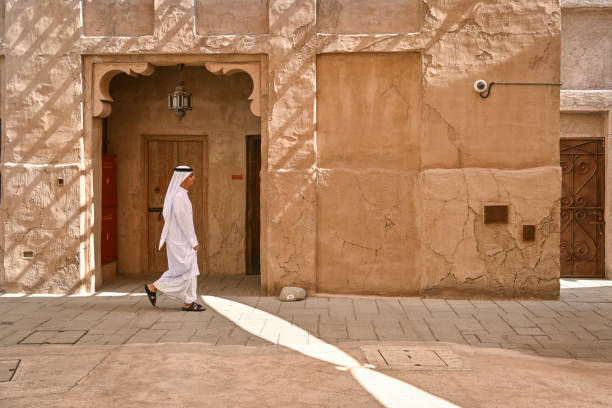 uomo arabo vestito di bianco thobe cammina accanto a una vecchia casa in una strada di al fahidi, un quartiere storico di dubai, emirati arabi uniti - old stone house foto e immagini stock