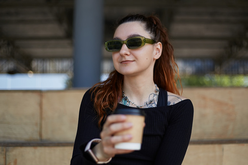Portrait of a young tattooed woman in sunglasses drinking coffee from a disposable paper cup