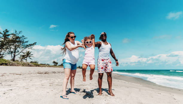 Multiracial family with pregnant mother father and toddler daughter walk and play on an idyllic beach together on vacation Beautiful young family walk on the beach together family beach vacations travel stock pictures, royalty-free photos & images