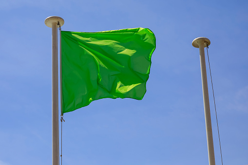 A green rectangular flag fluttering in the wind against a blue sky. The second flagpole right next to it is now empty, without a flag