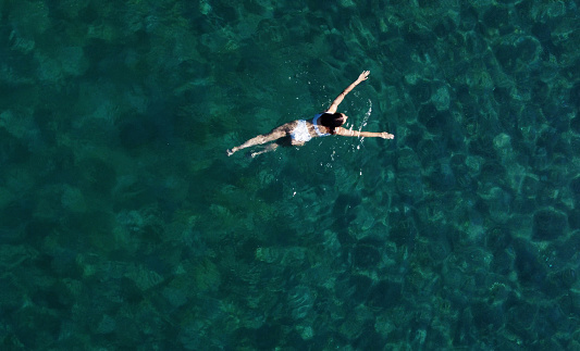 Above view of woman swimming at sea. Aerial top view of young girl in white swimsuit floating on water surface in crystal clear turquoise sea. Vacation at Paradise. Ocean relax, travel and vacation.