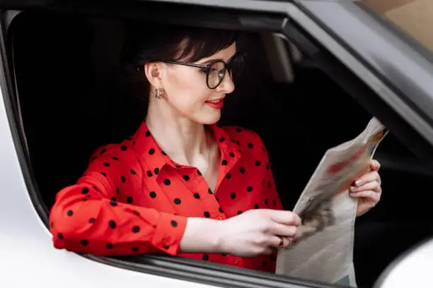 Young beautiful businesswoman with red lipstick in glasses and red polkadots blouse sitting in her luxurious car looking in camera and holding a newspaper. Business concept.