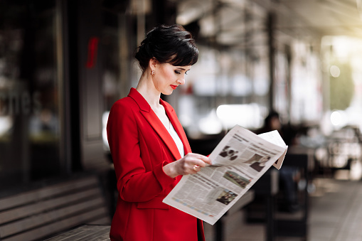 Stylish beautiful brunette woman wearing red jacket holds newspaper and reads news in the morning outdoors by the office centre. Elegant businesswoman has break for new information.