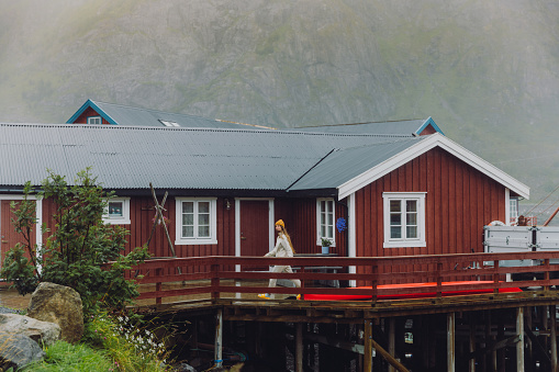 Side view of female in yellow hat walking by the old-fashioned fisherman's house to the seashore during summer rainy day on Lofoten, Northern Norway