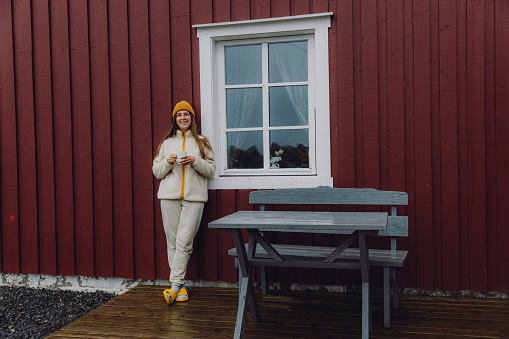 Female in yellow hat staying by the old-fashioned fisherman's house drinking coffee during summer rainy day on Lofoten, Northern Norway