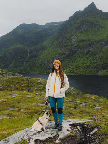 Smiling female with backpack traveler with long hai  in yellow hat hiking with cute pug in the mountains with lake view during summertime on Lofoten, Nordland, Northern Norway
