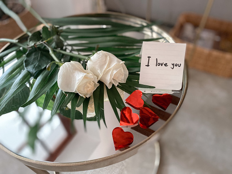 Still life with a white roses on a glass table and with the message ‘I love you’ handwritten on a piece of paper