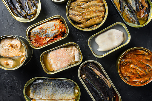 A group of aluminium cans canned with different types of fish and seafood, top view over black textured stone  background.