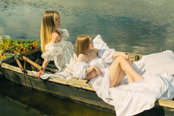deux femmes blondes en robe blanche profitant de vacances sur le lac. amitié féminine. draps blancs dans un vieux bateau de pêche avec rame. draps. détente estivale - deck chair photos et images de collection