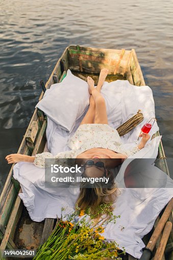 istock Blonde woman in white dress enjoying vacation on the lake. White linens in old fishing boat with oar. Bed sheets. Summer relaxation 1494042501