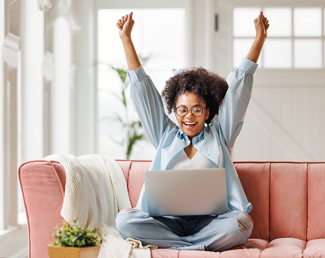 beautiful young smiling african american woman smiling and rejoices in victory   sitting on sofa and working at laptop screen after finishing project  in home office